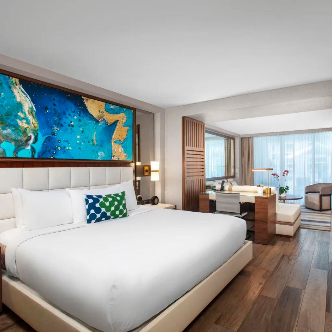 Hotel room with large bed and abstract painting above it. Beside the bed is a desk with curtained windows.